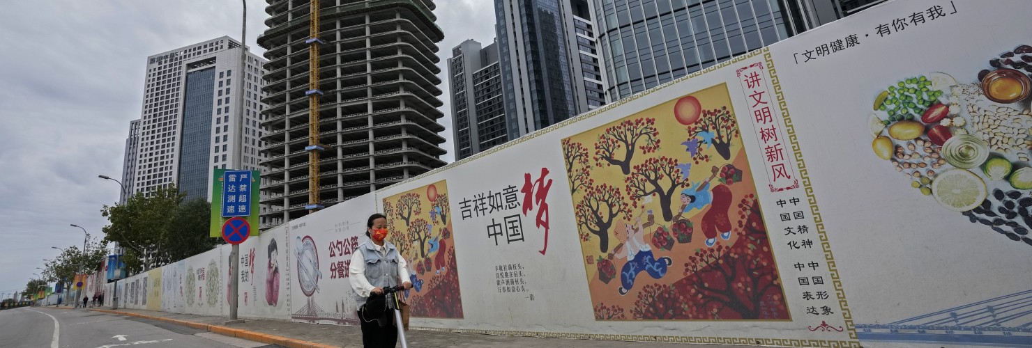 A girl wearing a face mask to help protect from the coronavirus rides a scooter past Chinese government's propaganda "China Dream" billboard on display along a commercial office buildings under construction in Tongzhou, outskirts of Beijing.