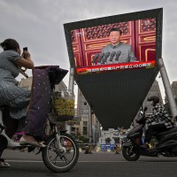 A woman on her electric-powered scooter films a large video screen outside a shopping mall showing Chinese President Xi Jinping speaking during an event to commemorate the 100th anniversary of China's Communist Party at Tiananmen Square in Beijing