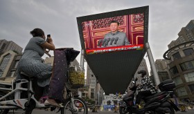 A woman on her electric-powered scooter films a large video screen outside a shopping mall showing Chinese President Xi Jinping speaking during an event to commemorate the 100th anniversary of China's Communist Party at Tiananmen Square in Beijing